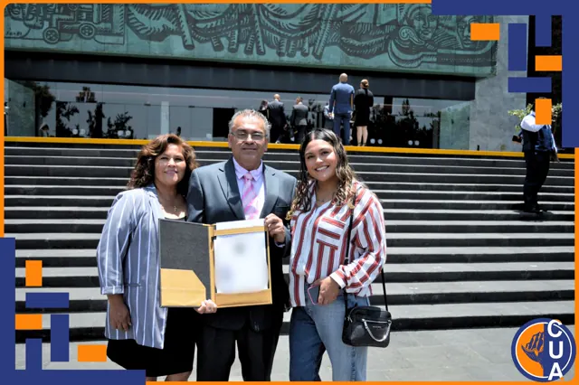 Egresado sonriente con traje y corbata rosa muestra su título acompañado por dos mujeres.