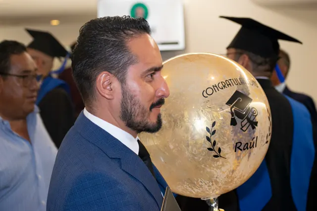 Egresado con traje azul sostiene un globo dorado de felicitación entre otros graduados.