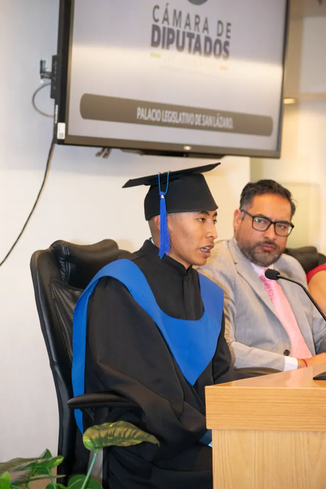 Graduado con toga negra y estola azul se dirige al público desde un atril.