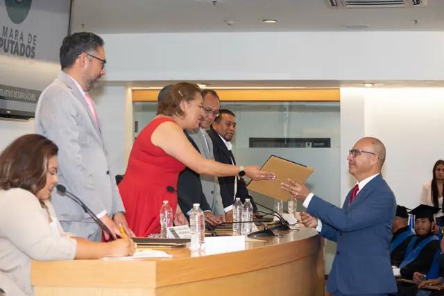 Egresado con traje azul y corbata roja recibe su carpeta de titulación.
