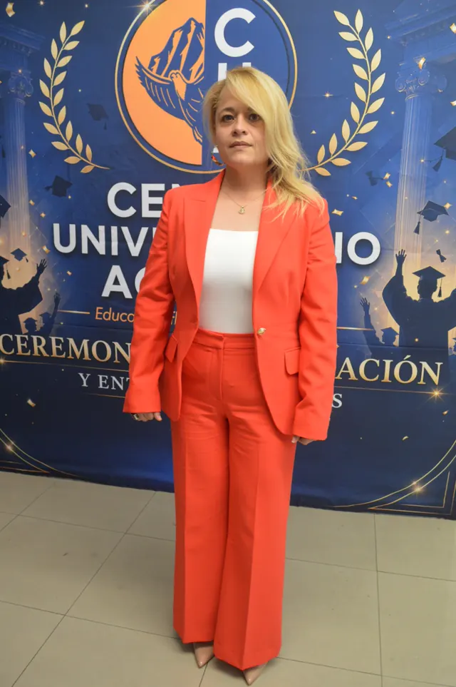 Participante con traje color coral posa frente al fondo de la ceremonia de graduación.