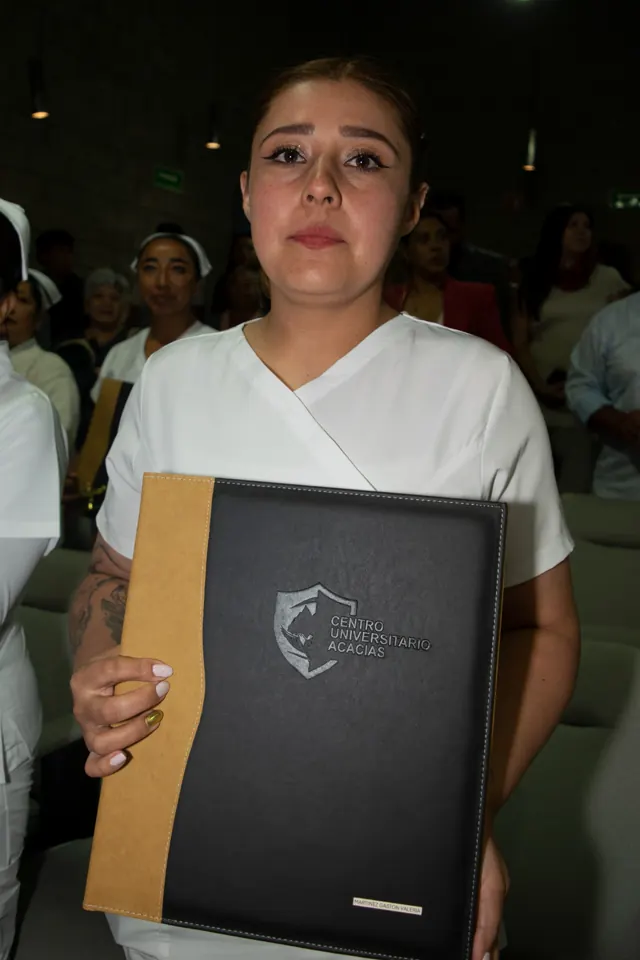 Egresada con uniforme blanco muestra su carpeta de graduación. Graduación del 14 de junio de 2025.